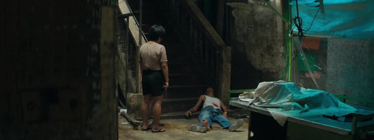 Person stands wiping tears looking down at bleeding man lying at food of stairs on floor
