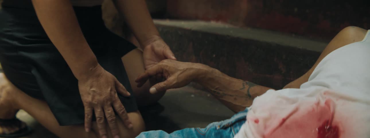 A woman kneeling down and holding the hand of a man laying in the bottom of the staircase bleeding blood