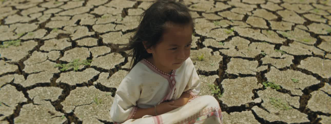 A young girl in a white dress squats on the cracked ground at a canyon