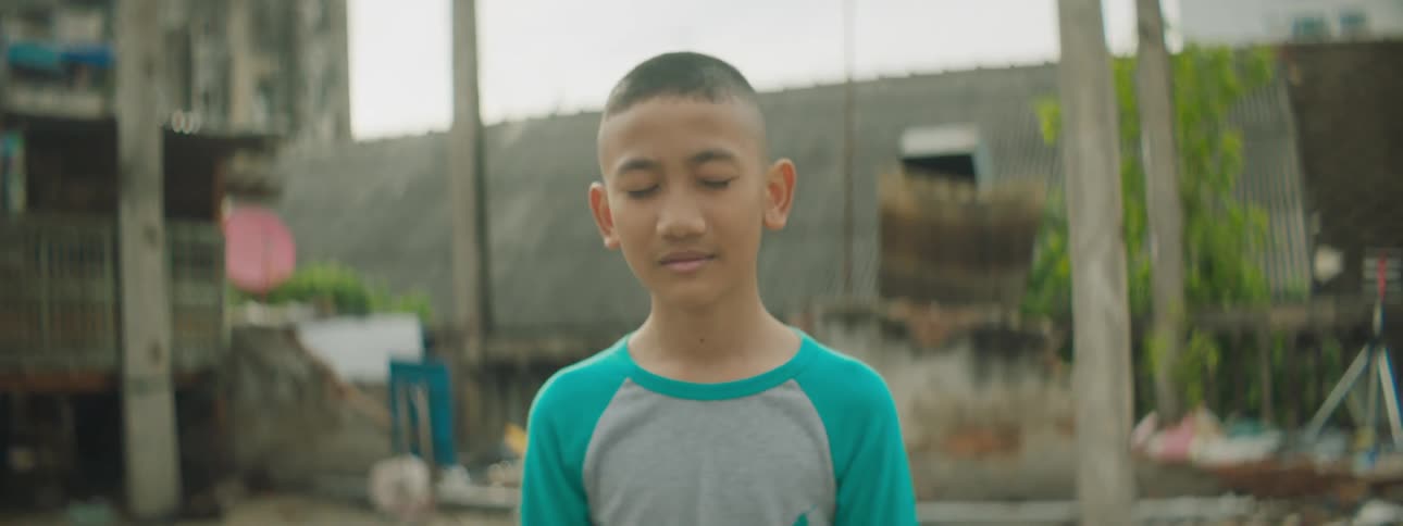 Young boy wearing stands outside closes eyes and smiles in front of wooden poles and some fencing with plants stuff and tall buildings in the background