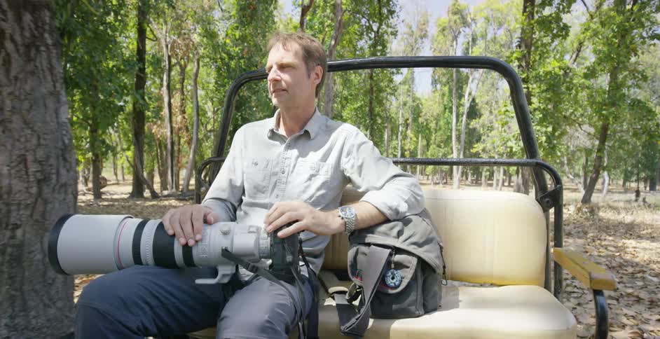 A man rides on the back of a vehicle in the woods carrying a large camera