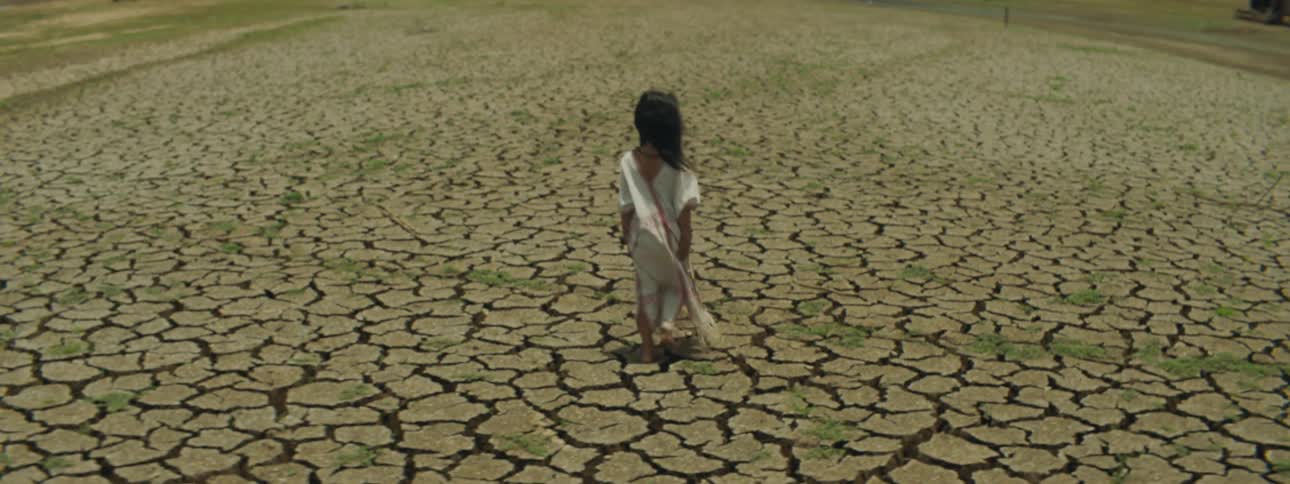 A young girl in a long white dress walks on cracked ground