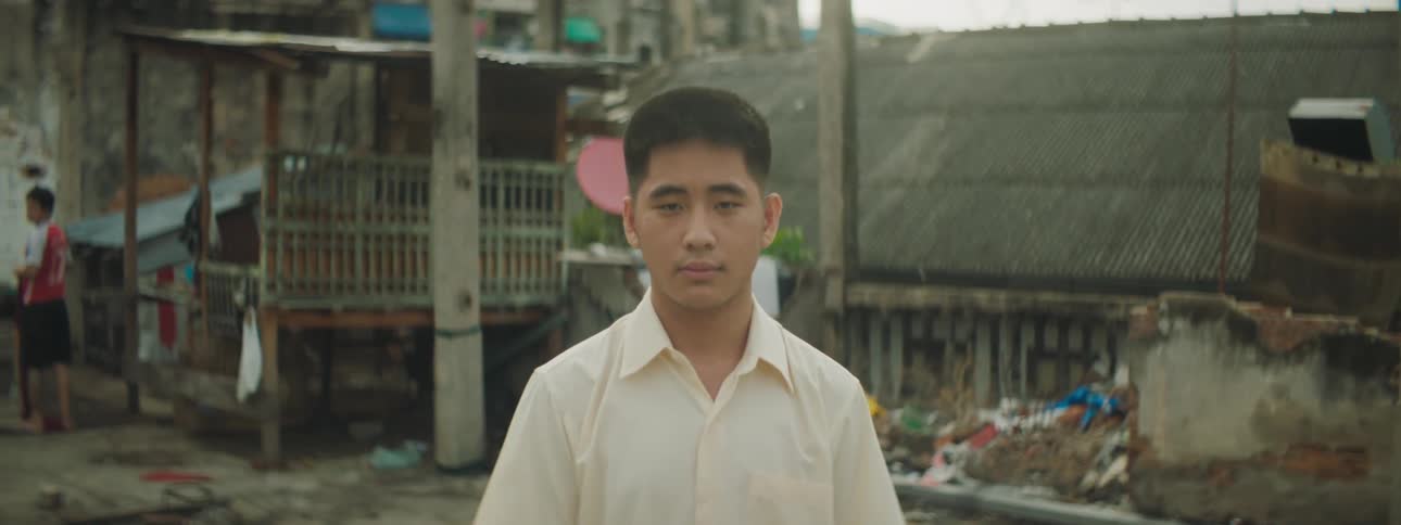 A young man stares into the camera while standing outdoors in the slums during the day
