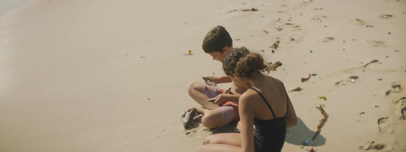 A boy and a girl sitting on the sand on the beach holding clams