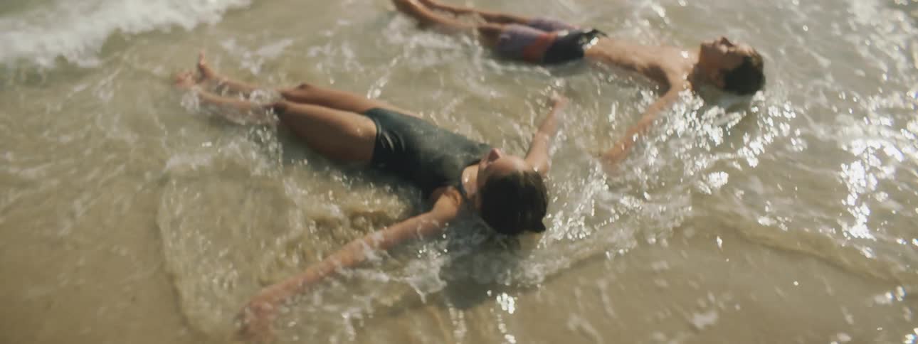 A girl and a boy laying on the beach with the waves splashing on their body