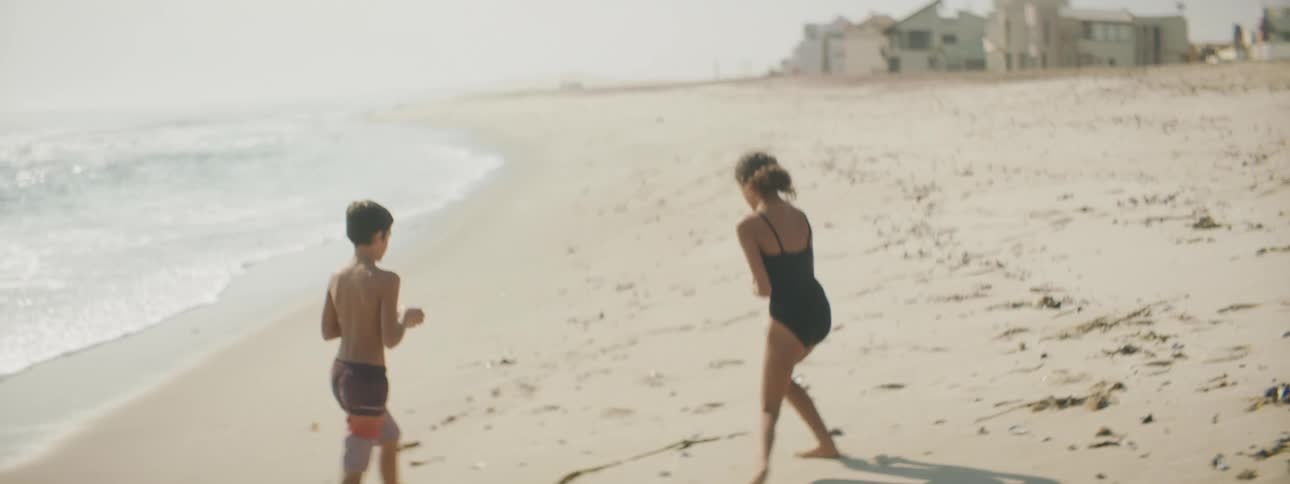 Boy and girl walk along beach near buildings collecting shells