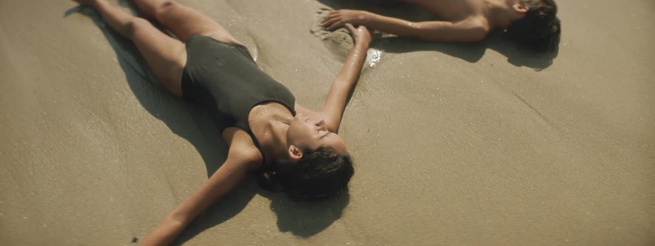 An ocean wave crashing over a brother and sister lying on a sandy beach in swimsuits