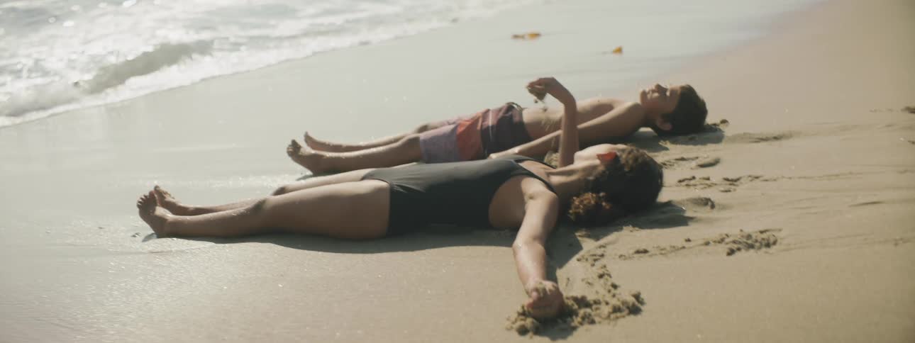A boy and girl lay on the beach at the ocean edge and the waves come crashing up to them