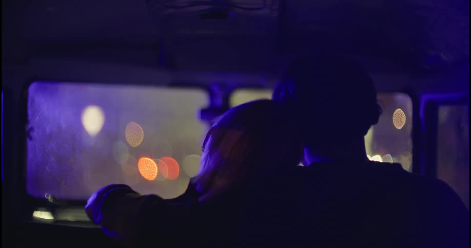 A young couple  cuddle in a car as red and blue emergency vehicle lights flash nearby