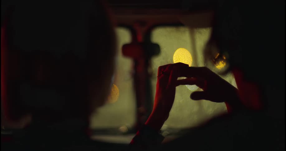 Two people are sitting inside an automobile and hold hands as the light from outside reflects on the windshield