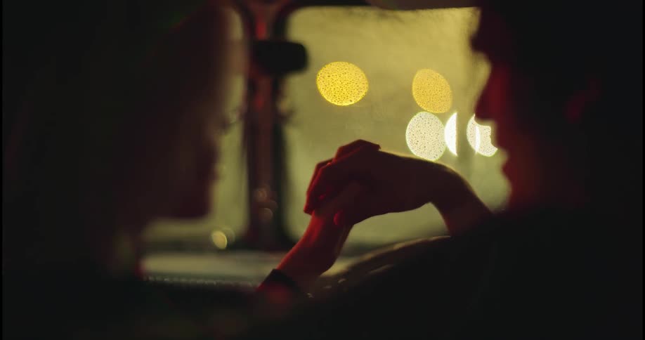 Two people are sitting inside an automobile and hold hands as the light from outside reflects on the windshield