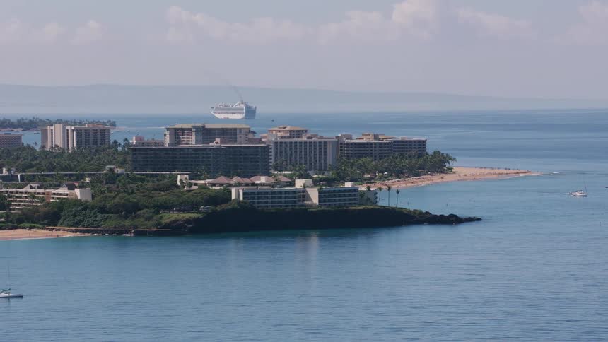 Buildings are along the coast as people are on the beach and a large cruise ship sails among smaller boats in the water as the waves roll into shore