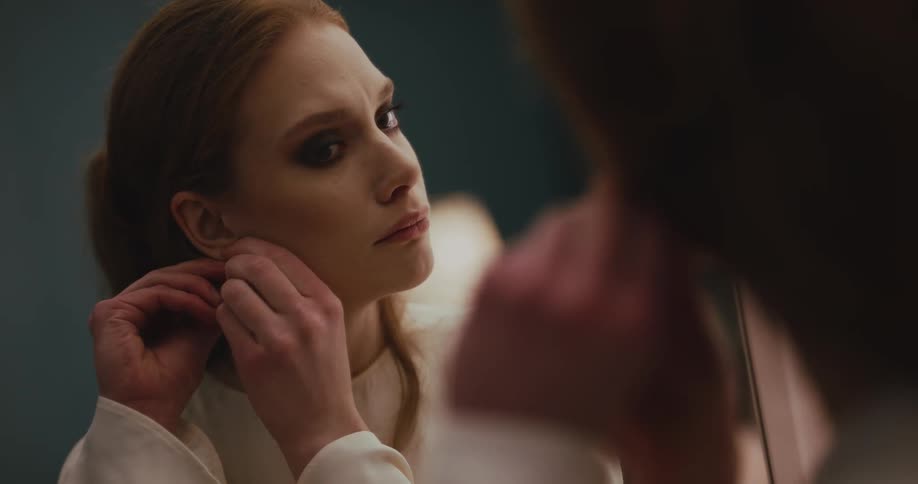 A woman puts on a pair of earrings while looking into a mirror with a distressed look on her face