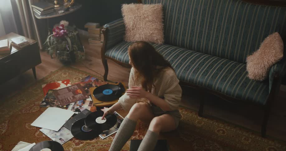 A woman sitting on the floor of her living room looks through a collection of vinyl records while enjoying the music