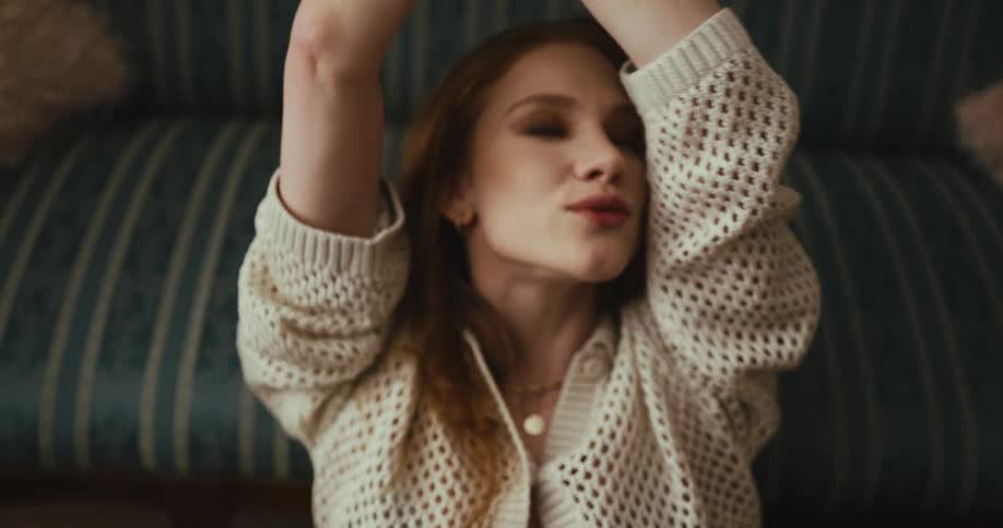 A young woman leans against a couch at home and raises her arms above her head and moves her head about and smiles