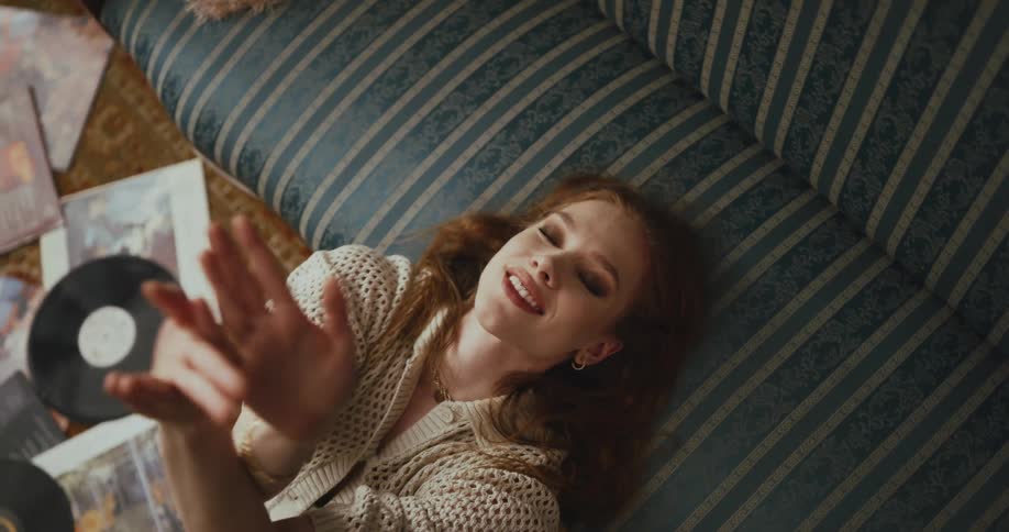 A woman happily waves her arms while she sits on the floor of her living room and listens to music