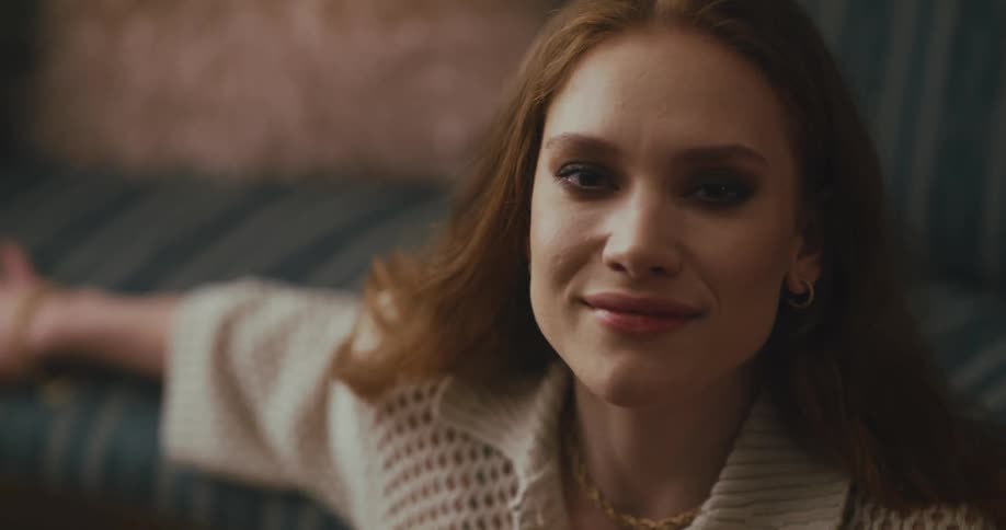 Redheaded woman sits on floor near couch swaying and smirking