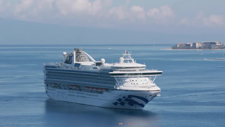 Small boats sail near a large cruise ship filled with people as it passes by land