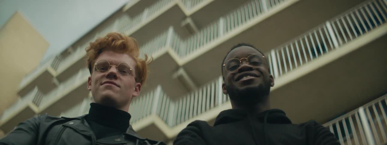 A pair of young men standing in front of a multistory building look downwards to smile into the camera