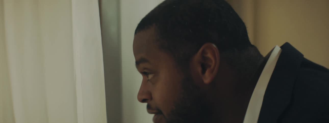A man in a room looking out of a window with blinds and smiling