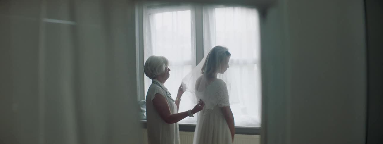 A woman turns to look at the camera peaking at her through the doorway as she has her wedding veil adjusted by her mother