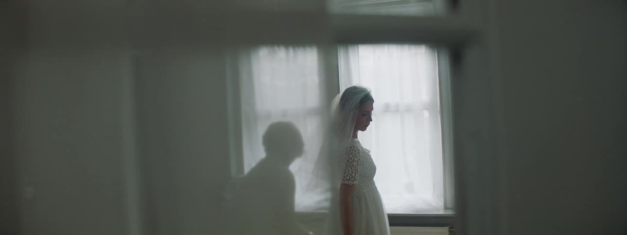 Elderly woman helps a younger woman with her wedding gown and veil as they stand in front of a window