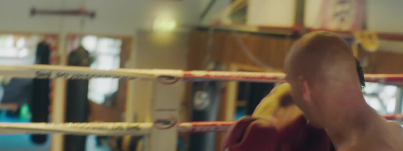 Two men wearing shorts and boxing gloves fight in a boxing ring the black guy knocks the white gut out behind them are some posters on the wall and sunlight outside the glass door
