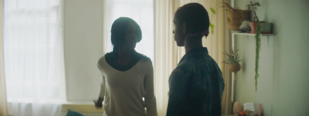 A boy and a woman in a room as the woman walks up and grabs an object from the boys hand with windows and curtains in the background