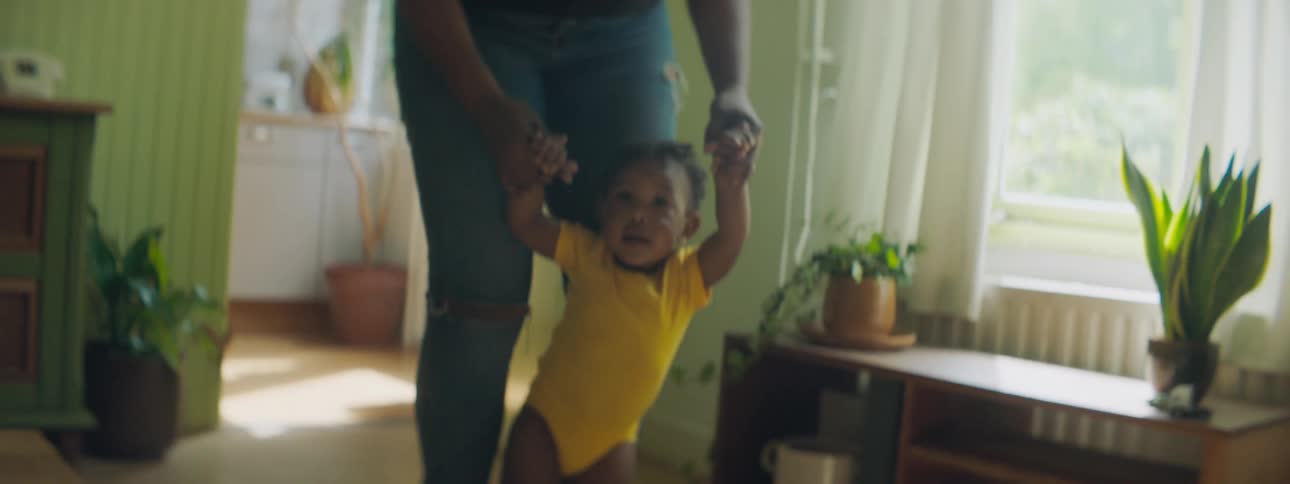 Mother helps toddling child walk through home past window and furniture