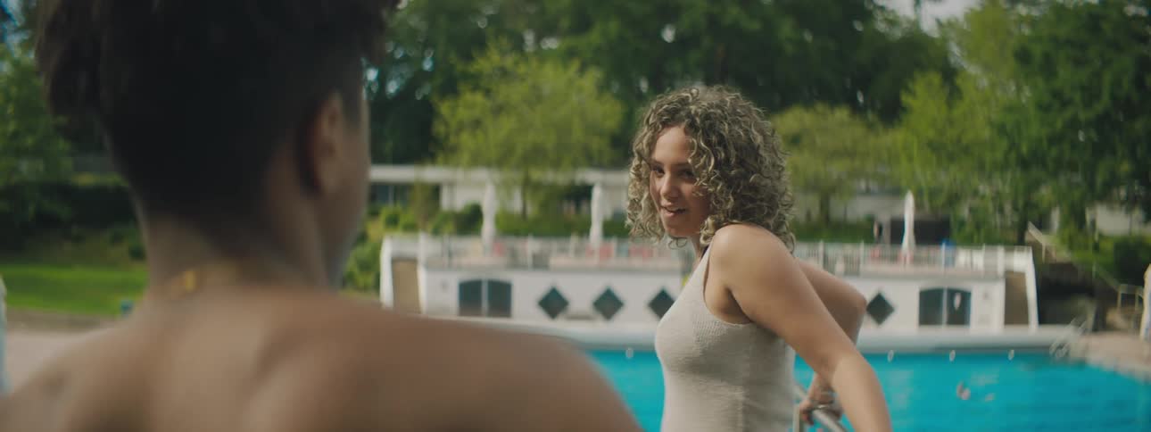 A woman flirtatiously looks at a man as they stand beside an outdoor pool