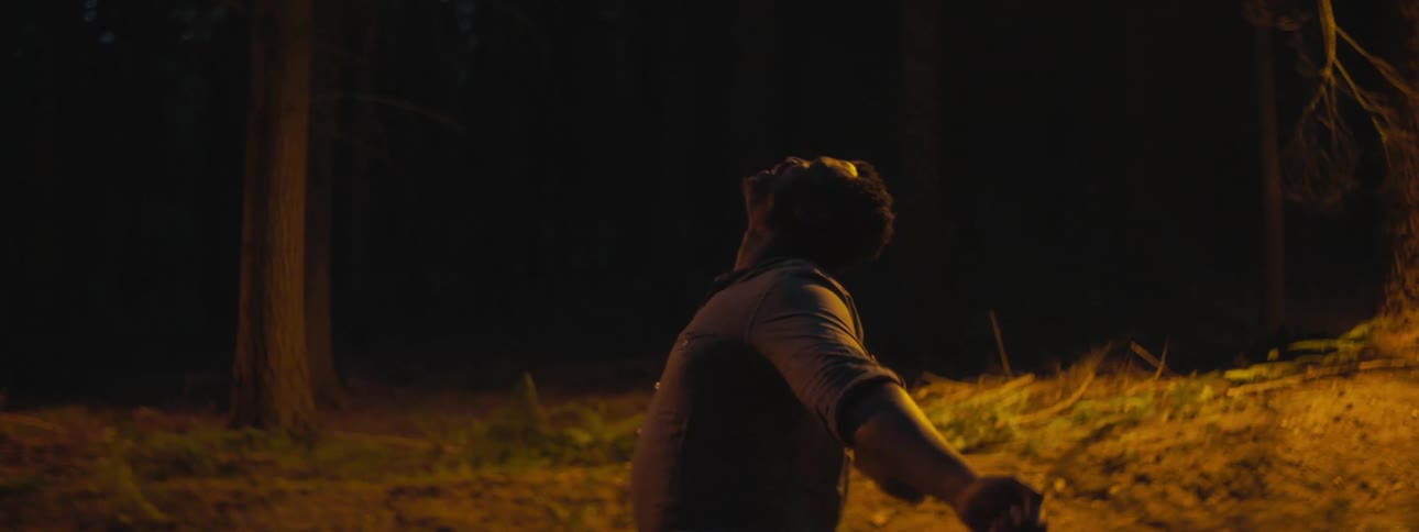 A man in a forest area stretches his arms to his side and then flops his head over and rests his hands on his knees