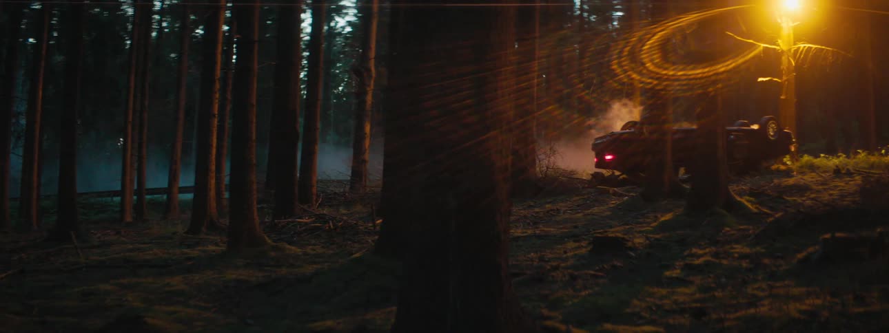 Man with a shirt walks up to a car crash upside down in the woods at dusk