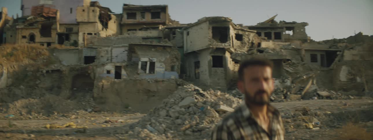 Man wearing a plaid shirt stands near piled rubble with ruined damaged buildings on hill in background