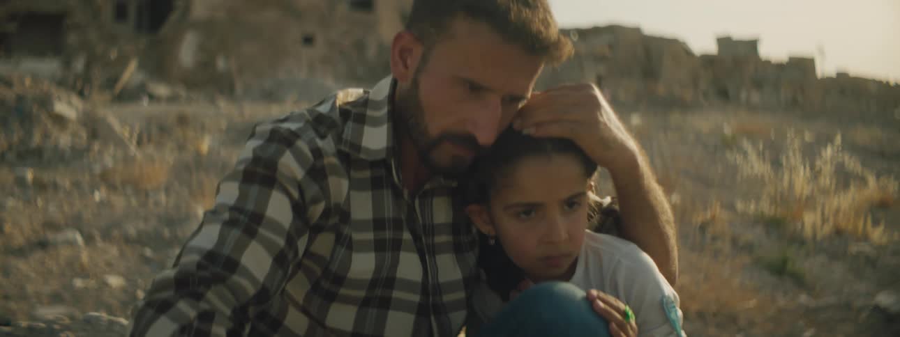 Father hold daughter close sitting on ground near ruined buildings