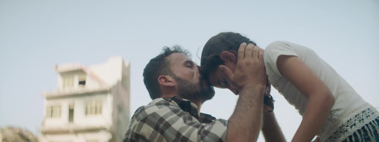 Father kisses daughter on forehead with ruined buildings and rubble nearby