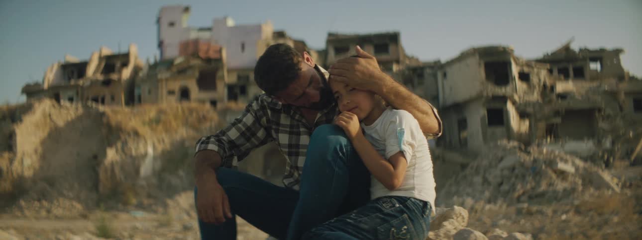 Father hugs daughter sitting on rubble in front of ruined town