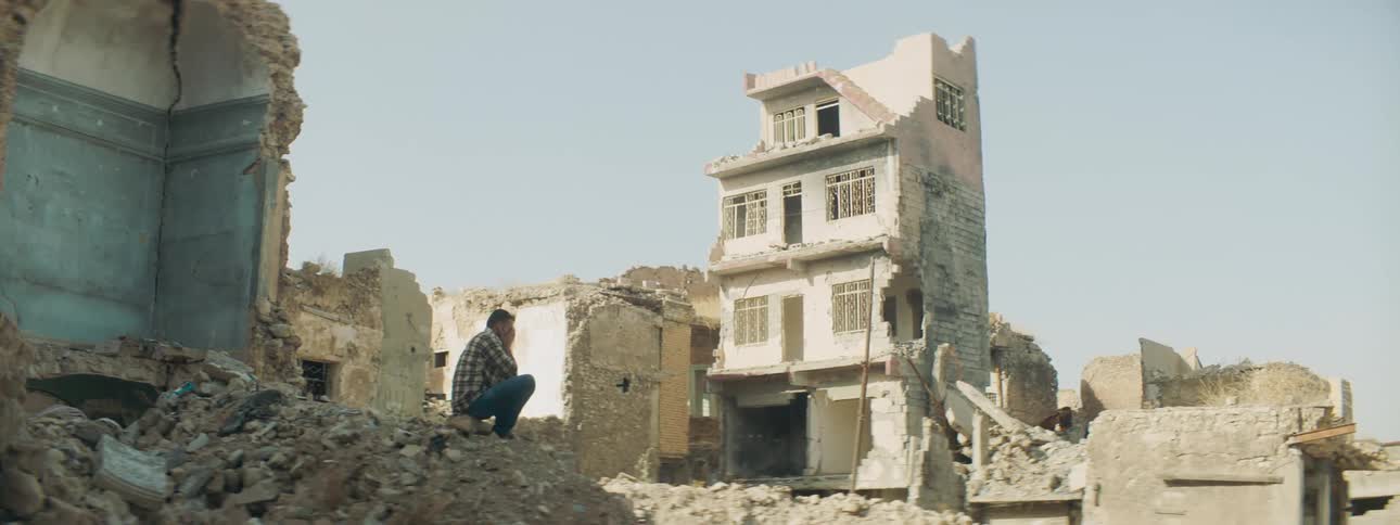 A man sits in rubble in a village with buildings falling apart