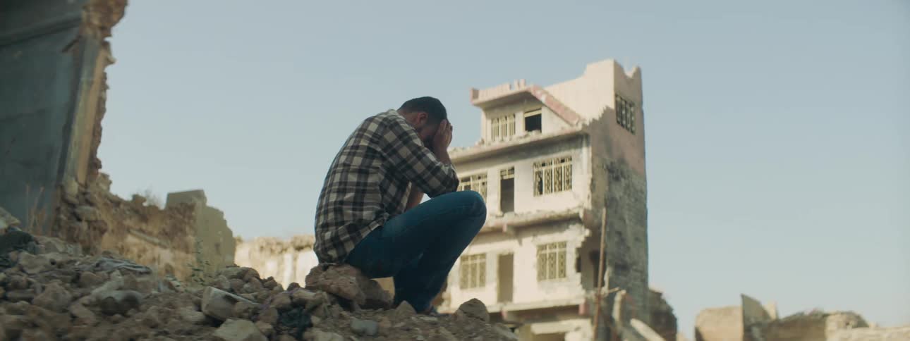 A man sitting on rubble with dilapidated buildings the sky and clouds in the background