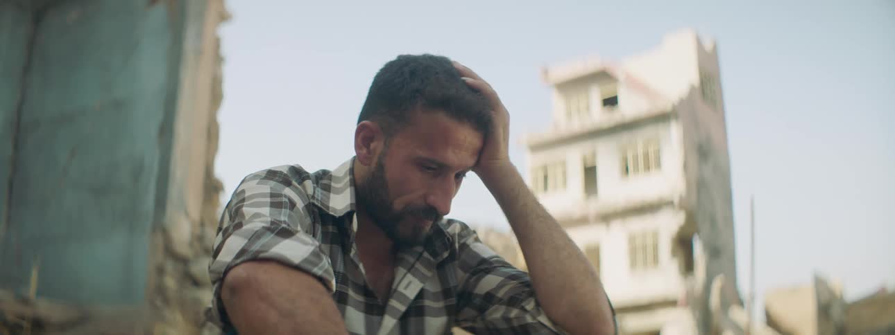 A man sits with his head in his hand in front of a building that is falling apart in a village