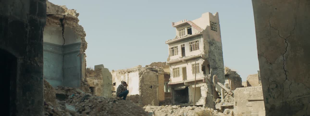 A man sitting down on rubble with dilapidated buildings and the sky and clouds in the background