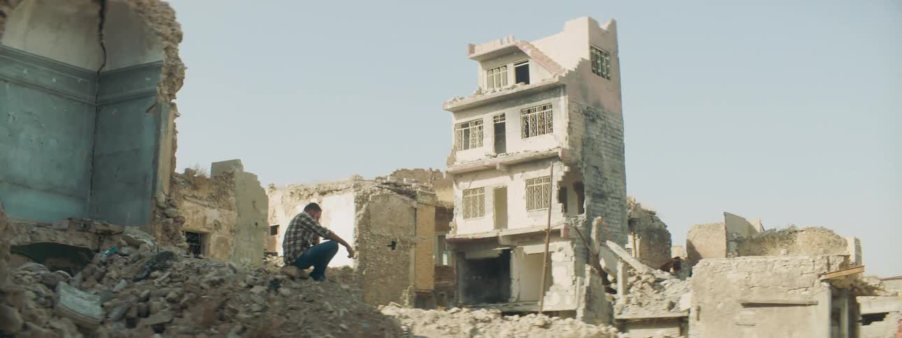 An upset man sits on a pile of rubble surveying the destroyed buildings around him