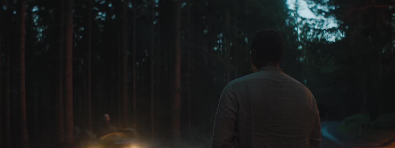 A man looks at another person standing in front of a car with headlights on in a wooded area