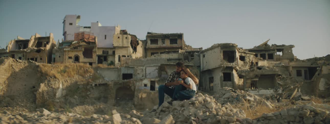A man and a girl sitting on rubble hugging with dilapidated buildings the sky and clouds in the background