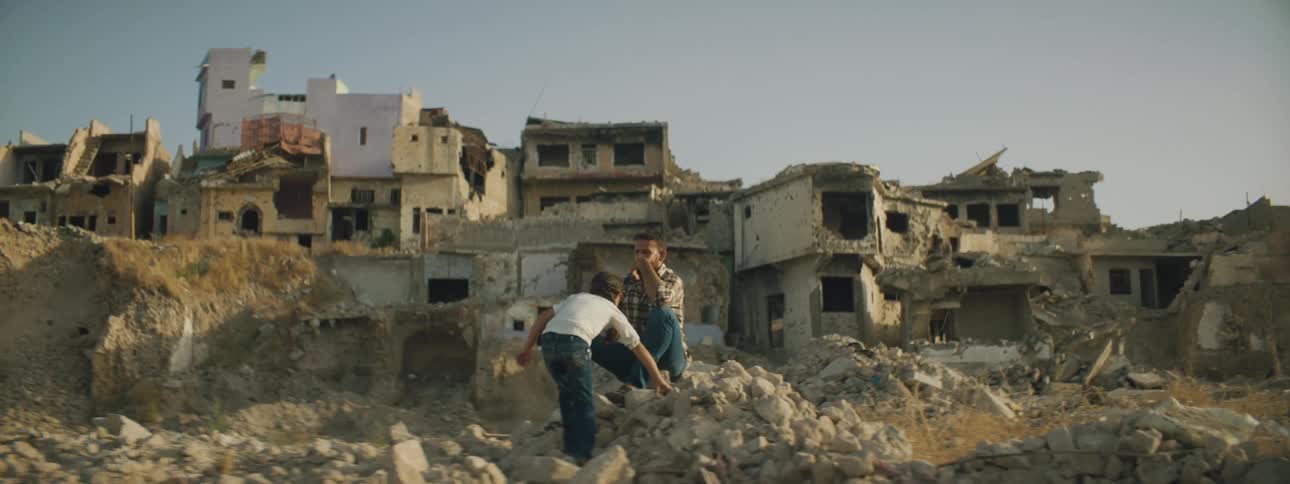 A girl carefully walks across rocks to approach a man sitting on a pile of rubble in front of a row of ruined buildings