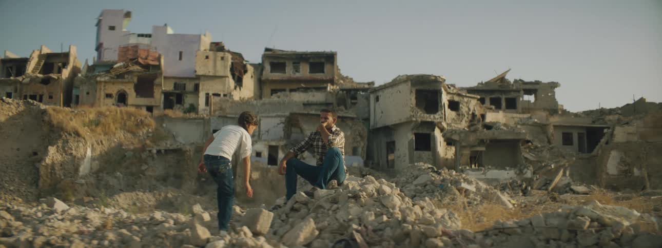 Man sitting on a pile of rocks from destroyed buildings with child walking towards him
