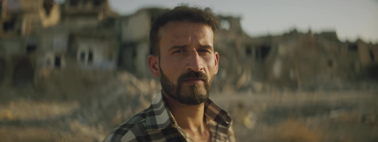 Man with beard and mustache wearing plaid shirt stand near pile of rubble with ruined buildings in background
