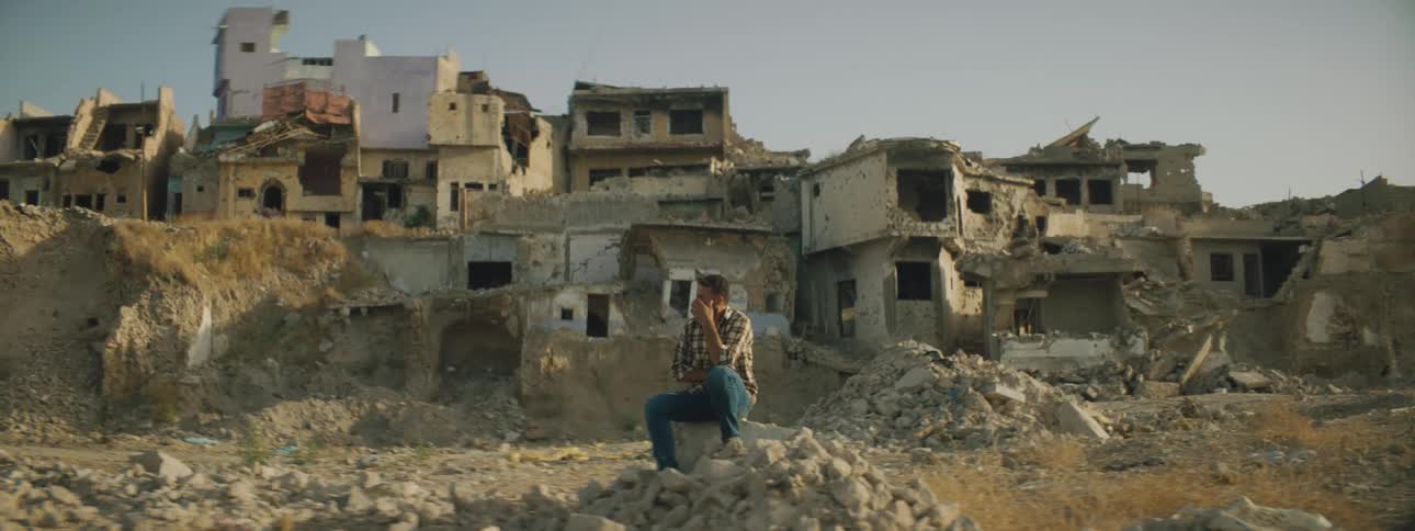 Man sits on block in front of rubble strewn area of destroyed buildings