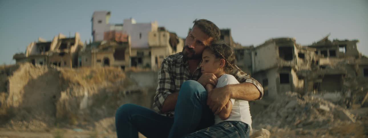 A man hugs his daughter and points ahead of him as they sit in front of buildings that are falling apart