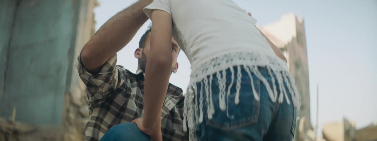 An upset man sitting in front of the ruins of a building lifts his head as a girl approaches and kisses her forehead
