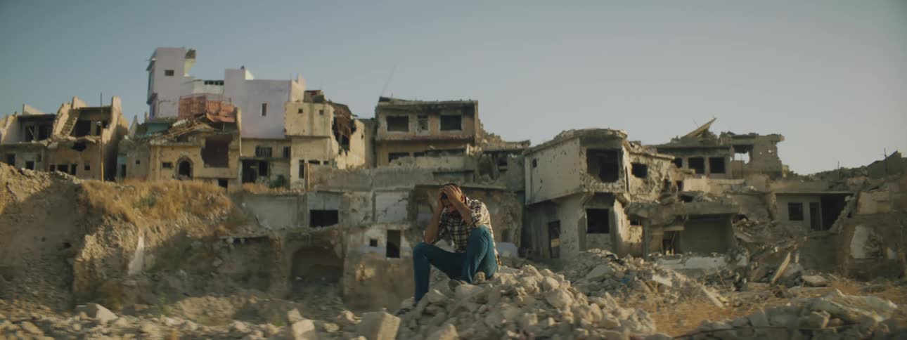 A man with his head in his hands sits on a pile of stones in front of buildings that have been destroyed